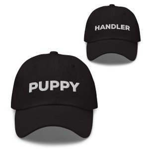 May include: Two black baseball caps with white embroidered text. One cap says "PUPPY" and the other says "HANDLER".