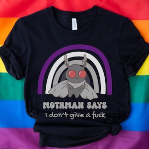 Könnte beinhalten: Schwarzes T-Shirt mit einer Cartoon-Motten-Illustration, roten Augen, Geweih und einem Umhang, vor einem Regenbogen-Design. Der Text "Mothman Says I don't give a fuck" ist darunter gedruckt. Das Shirt wird auf einer Regenbogenflagge präsentiert.