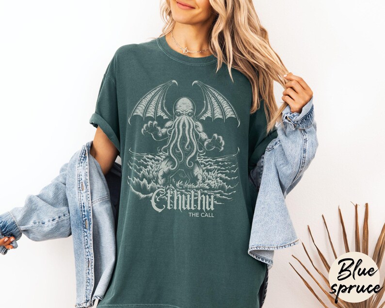 May include: A teal green t-shirt with a graphic print of Cthulhu, a fictional tentacled monster from the works of H.P. Lovecraft. The text "Cthulhu The Call" is printed below the graphic.