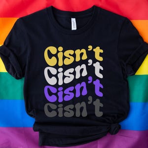 Może przedstawiać: Czarny T-shirt z napisem "Cis'n't Cis'n't Cis'n't Cis'n't" w tęczowych kolorach. Koszulka leży na tęczowej fladze dumy.