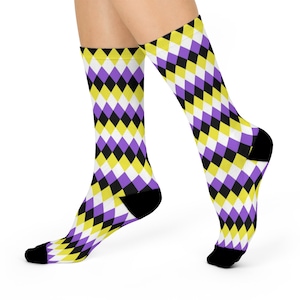 Peut inclure: Paire de chaussettes avec un motif de losanges répété en jaune, violet, noir et blanc. Les chaussettes ont des talons et des orteils noirs. Les chaussettes sont sur les pieds d'une personne.
