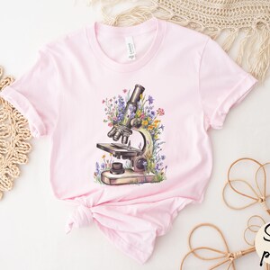 May include: A light pink t-shirt featuring a watercolor illustration of a microscope surrounded by colorful flowers. The shirt has a classic crew neck and short sleeves. The words "Soft pink" are in a circle on the bottom right.