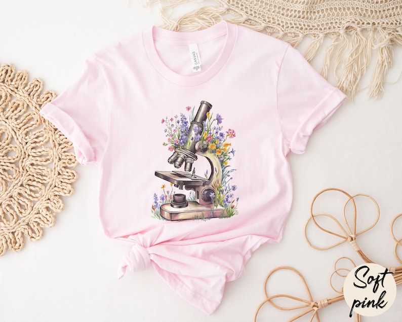 May include: A light pink t-shirt featuring a watercolor illustration of a microscope surrounded by colorful flowers. The shirt has a classic crew neck and short sleeves. The words "Soft pink" are in a circle on the bottom right.