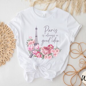 May include: White t-shirt with a design featuring the Eiffel Tower and pink and purple flowers. The text "Paris is always a good idea" is printed above the design. The shirt is tied at the waist.