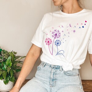 May include: White t-shirt featuring a graphic of three pink, purple, and blue dandelions with seeds blowing in the wind. Two small pink and blue hearts are in the top right corner of the design.