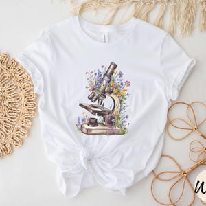 May include: White t-shirt featuring a watercolor illustration of a vintage microscope surrounded by colorful wildflowers. The design includes details like the eyepiece, objective lenses, and stage of the microscope. The shirt is made of a soft, white fabric.