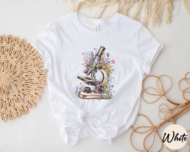 May include: White t-shirt featuring a watercolor illustration of a vintage microscope surrounded by colorful wildflowers. The design includes details like the eyepiece, objective lenses, and stage of the microscope. The shirt is made of a soft, white fabric.