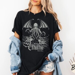 May include: Black t-shirt with a graphic print of Cthulhu, a fictional creature with tentacles and wings, rising from the sea. The text "Cthulhu The Call" is printed below the image.