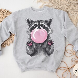 May include: A gray sweatshirt with a cartoon raccoon wearing a pink bubble. The raccoon is looking at the bubble and has a happy expression.
