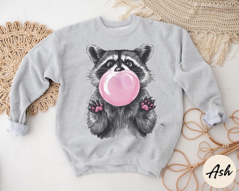 May include: A gray sweatshirt with a cartoon raccoon wearing a pink bubble. The raccoon is looking at the bubble and has a happy expression.
