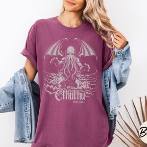 May include: A maroon t-shirt with a white graphic of Cthulhu, a fictional tentacled monster from the works of H.P. Lovecraft. The graphic includes the text "Cthulhu" and "The Call".