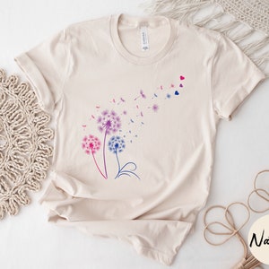 May include: A beige t-shirt featuring a graphic of three pink, blue, and purple dandelions with seeds blowing in the wind. The dandelions are outlined in black and have a few small red hearts scattered around them.
