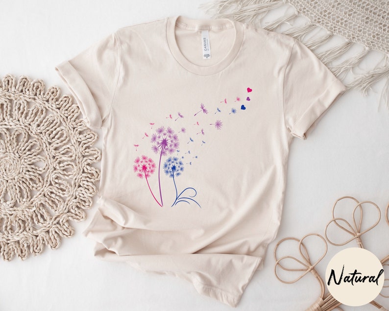 May include: A beige t-shirt featuring a graphic of three pink, blue, and purple dandelions with seeds blowing in the wind. The dandelions are outlined in black and have a few small red hearts scattered around them.