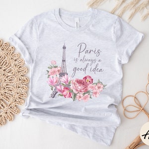 May include: Pale grey t-shirt with a design featuring the Eiffel Tower, pink flowers, and the phrase "Paris is always a good idea." The floral arrangement is in a watercolour style. The shirt is made of a soft material.