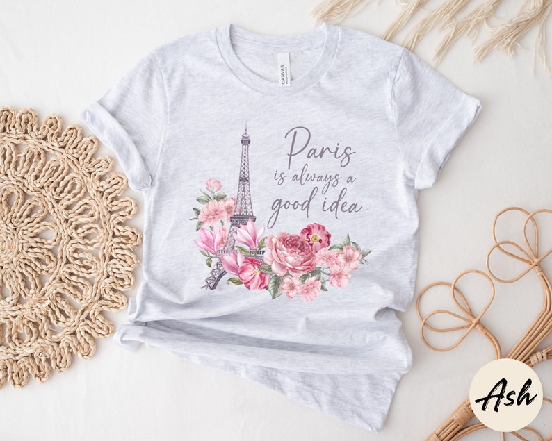 May include: Pale grey t-shirt with a design featuring the Eiffel Tower, pink flowers, and the phrase "Paris is always a good idea." The floral arrangement is in a watercolour style. The shirt is made of a soft material.