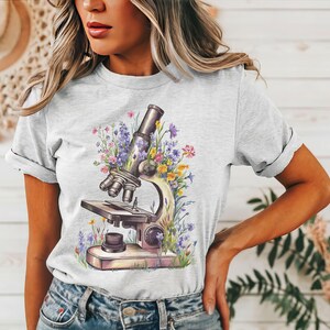 May include: Light gray t-shirt featuring a watercolor illustration of a microscope surrounded by colorful flowers. The design includes purple, yellow, and pink blooms, creating a botanical and scientific theme. The shirt has rolled-up sleeves and a casual fit.