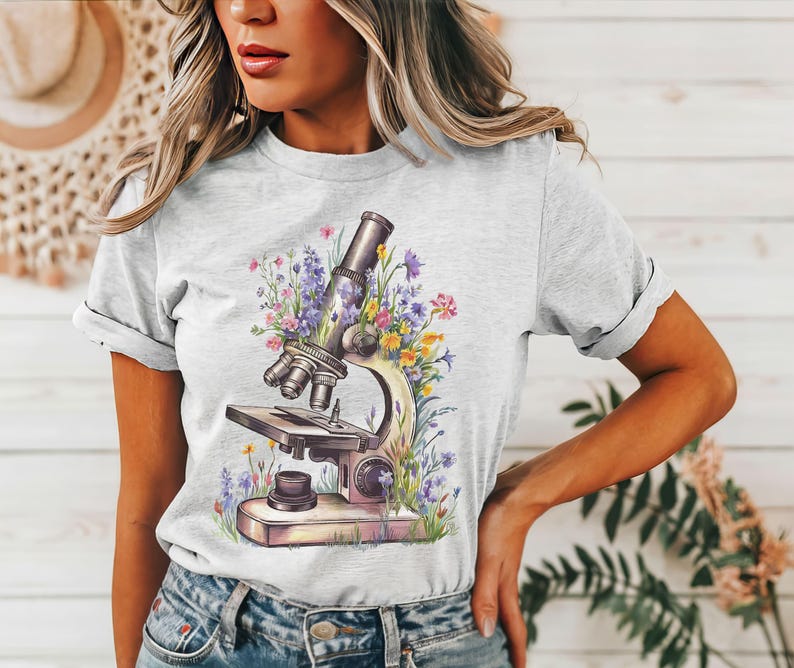 May include: Light gray t-shirt featuring a watercolor illustration of a microscope surrounded by colorful flowers. The design includes purple, yellow, and pink blooms, creating a botanical and scientific theme. The shirt has rolled-up sleeves and a casual fit.