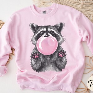 May include: Pink sweatshirt with a graphic of a raccoon blowing a bubble. The raccoon is black and gray with a pink bubble.