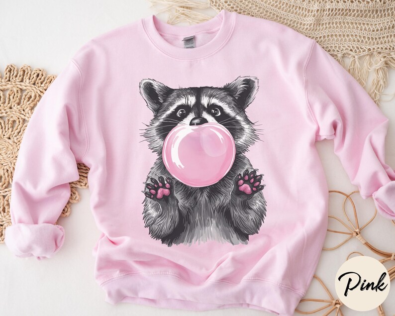 May include: Pink sweatshirt with a graphic of a raccoon blowing a bubble. The raccoon is black and gray with a pink bubble.