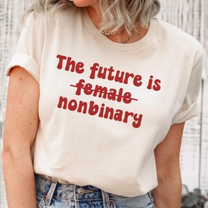 Może przedstawiać: Kremowa koszulka z czerwonym napisem "The future is nonbinary". Słowo "female" jest przekreślone. Koszulka ma podwinięte rękawy i jest noszona przez osobę. Osoba ma na sobie niebieskie dżinsy.