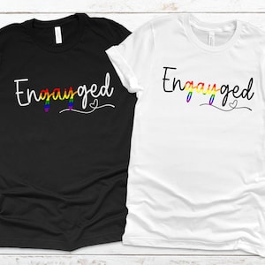 May include: Two t-shirts, one black and one white, with the word "Engayged" printed in white with a rainbow design. The word "Engayged" is stylized with a heart at the end.