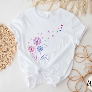 May include: White t-shirt featuring a design of three purple, pink, and blue dandelions with seeds blowing in the wind. The dandelions are outlined in black and have a few small red hearts scattered around them.
