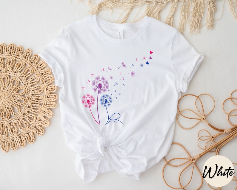 May include: White t-shirt featuring a design of three purple, pink, and blue dandelions with seeds blowing in the wind. The dandelions are outlined in black and have a few small red hearts scattered around them.