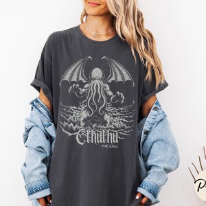 May include: A dark gray t-shirt with a graphic print of Cthulhu, a fictional monster from the works of H.P. Lovecraft. The text "Cthulhu The Call" is printed below the graphic.