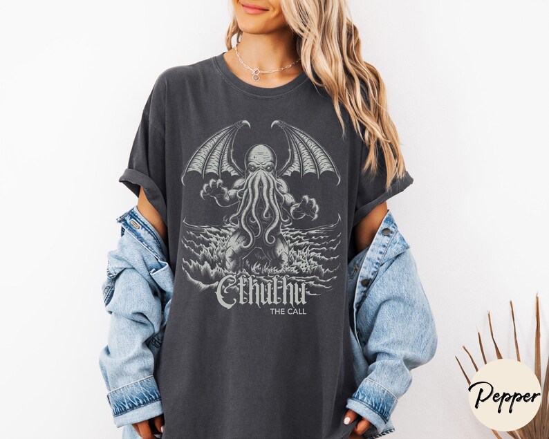 May include: A dark gray t-shirt with a graphic print of Cthulhu, a fictional monster from the works of H.P. Lovecraft. The text "Cthulhu The Call" is printed below the graphic.