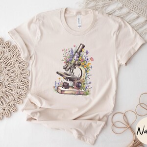 May include: Beige t-shirt featuring a watercolor illustration of a microscope surrounded by colorful wildflowers. The shirt has short sleeves and a crew neck. The word "Natural" is printed on a round sticker.