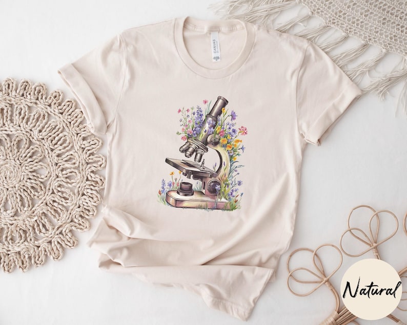 May include: Beige t-shirt featuring a watercolor illustration of a microscope surrounded by colorful wildflowers. The shirt has short sleeves and a crew neck. The word "Natural" is printed on a round sticker.