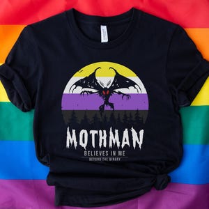 Könnte beinhalten: Schwarzes T-Shirt mit einer Grafik von Mothman vor einem Sonnenuntergang mit einem lila, weißen und gelben Streifen. Der Text "Mothman Believes in Me Beyond the Binary" ist unter der Grafik gedruckt.