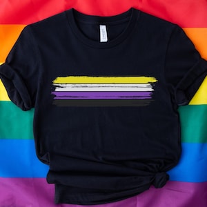 Może przedstawiać: Czarny t-shirt z namalowanym wzorem flagi dumy osób niebinarnych. Flaga ma żółte, białe, fioletowe i czarne pasy.