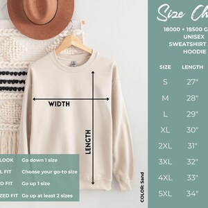 May include: Size chart for unisex sweatshirts and hoodies made by Gildan. The chart shows the length and width in inches for sizes S through 5XL. The chart is displayed on a light beige sweatshirt.