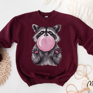 May include: A maroon sweatshirt with a graphic of a raccoon blowing a bubble. The raccoon is wearing a pink bubble and has a black nose and eyes.