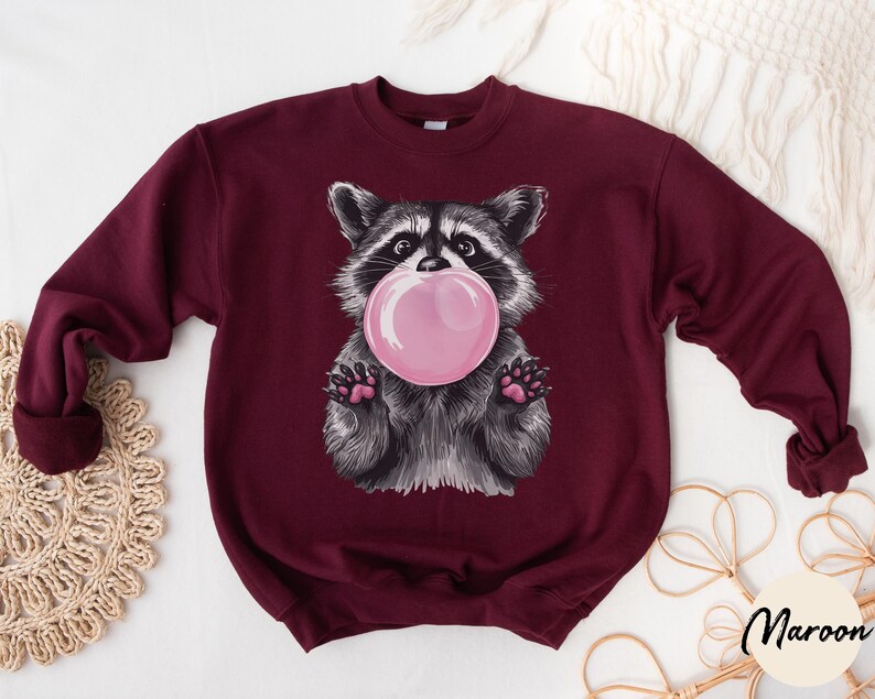 May include: A maroon sweatshirt with a graphic of a raccoon blowing a bubble. The raccoon is wearing a pink bubble and has a black nose and eyes.