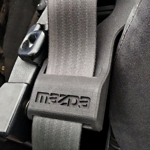 Może przedstawiać: Czarny pas bezpieczeństwa z wytłoczonym napisem "MAZDA" na klamrze. Pas bezpieczeństwa znajduje się w samochodzie.