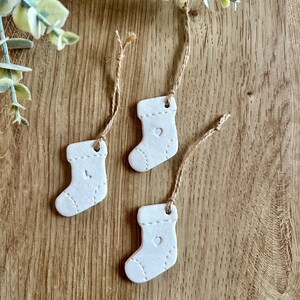 May include: Three white ceramic Christmas stocking ornaments with a heart design and twine for hanging.