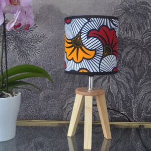 May include: A table lamp with a cylindrical fabric shade featuring a bold red, orange, and white graphic floral pattern. The lamp has a light-colored wooden tripod base with three angled legs and a small circular top platform supporting the shade.