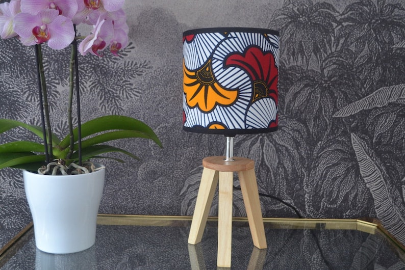 May include: A table lamp with a cylindrical fabric shade featuring a bold red, orange, and white graphic floral pattern. The lamp has a light-colored wooden tripod base with three angled legs and a small circular top platform supporting the shade.