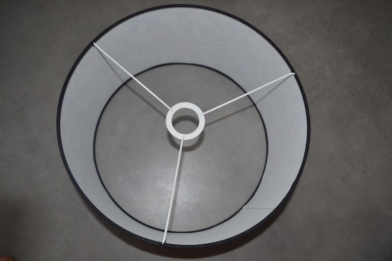 May include: A circular lamp shade featuring a black exterior and a white interior lining. The structure includes a white metal spider fitter with three spokes connecting to a central ring, designed for attachment to a lamp base or fixture.