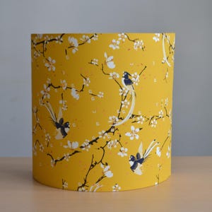 Puede incluir: Pantalla de lámpara cilíndrica con un fondo amarillo vibrante. Presenta un patrón repetido de flores blancas, ramas negras y pájaros estilizados. El diseño evoca un tema natural, adecuado para la decoración de interiores. El diámetro de la pantalla es de unos 20 cm.