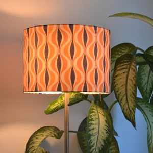 May include: A floor lamp with a cylindrical lampshade featuring a retro orange and black wavy pattern. The lamp is positioned next to a green leafy plant.