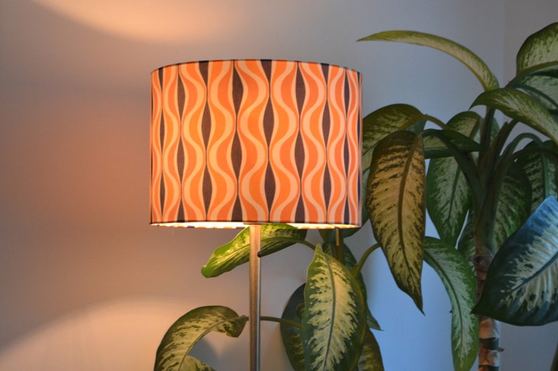 May include: A floor lamp with a cylindrical lampshade featuring a retro orange and black wavy pattern. The lamp is positioned next to a green leafy plant.