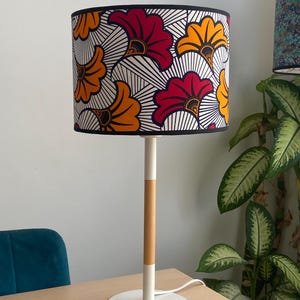 May include: A table lamp featuring a cylindrical lampshade with a vibrant pattern of red and orange fan-shaped floral motifs on a white background with black lines. The lamp has a slender white and light wood-toned stem and a round white base.