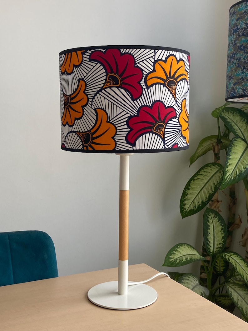 May include: A table lamp featuring a cylindrical lampshade with a vibrant pattern of red and orange fan-shaped floral motifs on a white background with black lines. The lamp has a slender white and light wood-toned stem and a round white base.