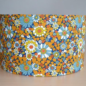 Peut inclure: Abat-jour cylindrique avec un motif floral vif. Le design présente des fleurs bleues, jaunes et blanches, ainsi que des cœurs orange, sur un fond bleu foncé. Un élément décoratif pour la maison.