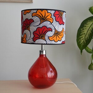 May include: A table lamp with a teardrop-shaped red glass base and a cylindrical lampshade. The lampshade features a repeating pattern of stylized orange and red flowers with black outlines on a white background, finished with a black trim.