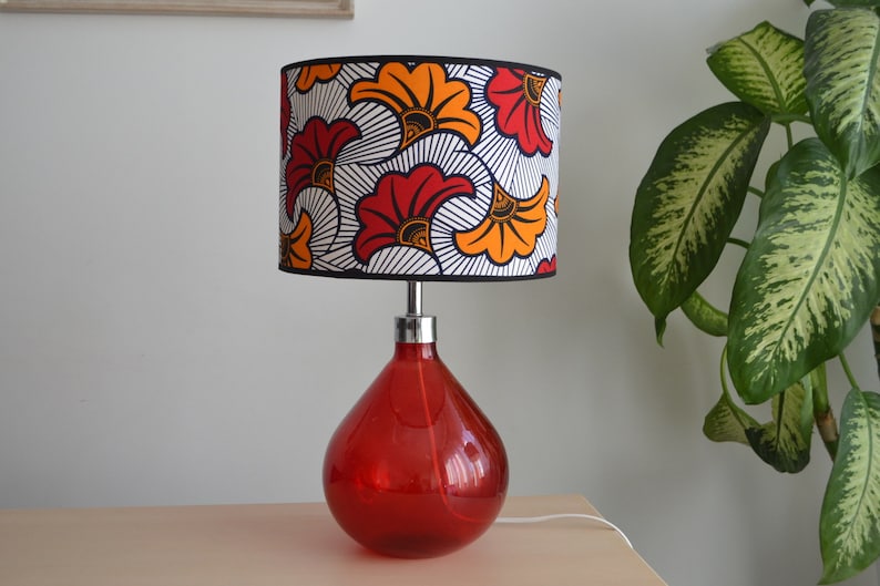 May include: A table lamp with a teardrop-shaped red glass base and a cylindrical lampshade. The lampshade features a repeating pattern of stylized orange and red flowers with black outlines on a white background, finished with a black trim.