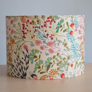 Op de afbeelding: Cilindrische lampenkap met een crèmekleurige achtergrond, versierd met een levendig bloemenpatroon. Het ontwerp bevat rode bessen, roze en gele bloemen en turquoise en groene bladeren. Een decoratief verlichtingselement.